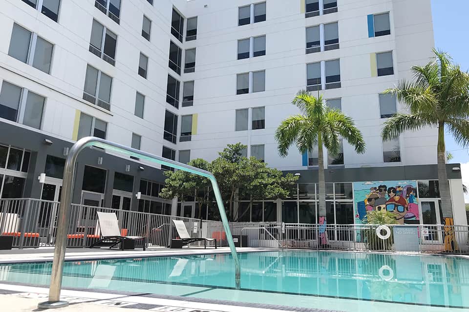 Modern white hotel building with turquoise pool, lounge chairs, and palm trees in courtyard