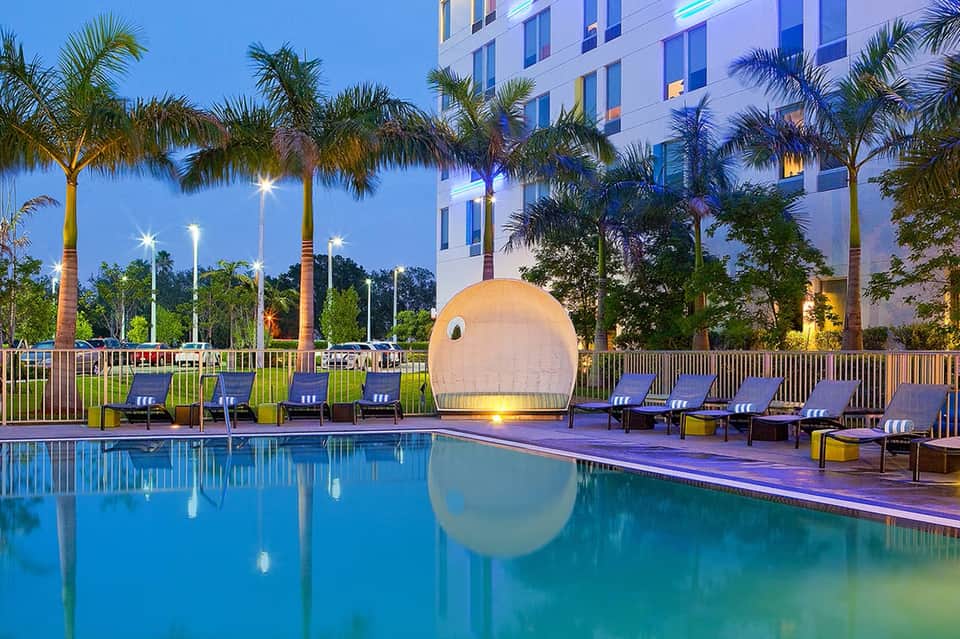 Modern hotel pool at dusk with lounge chairs, palm trees, and illuminated white building with distinctive dome structure