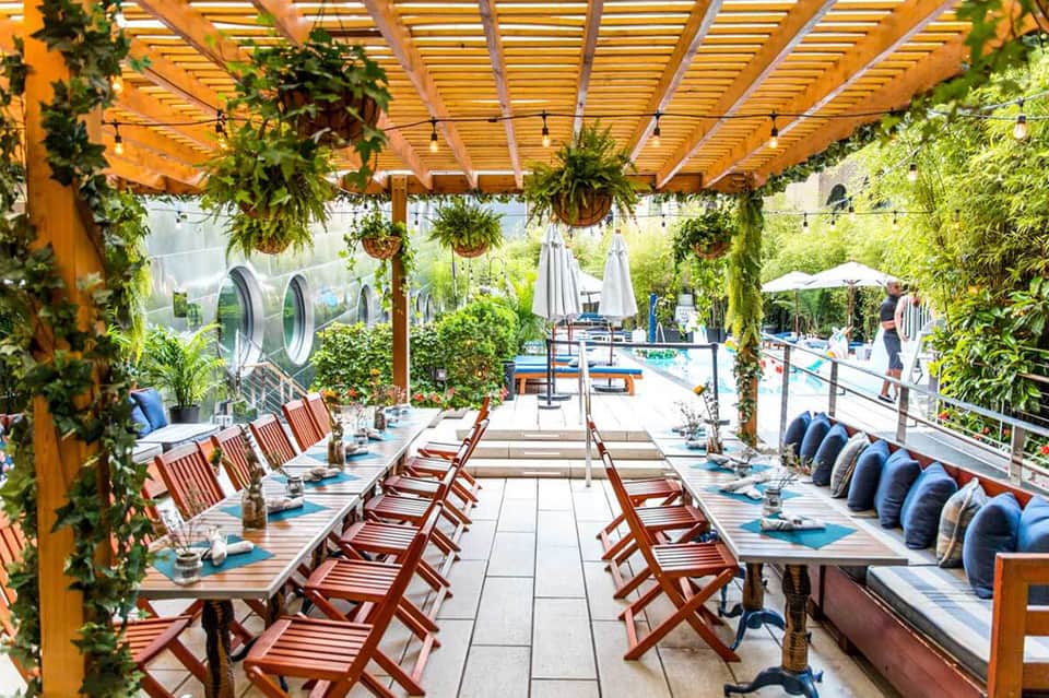Outdoor dining area with wooden pergola, hanging plants, and red chairs overlooking a resort pool