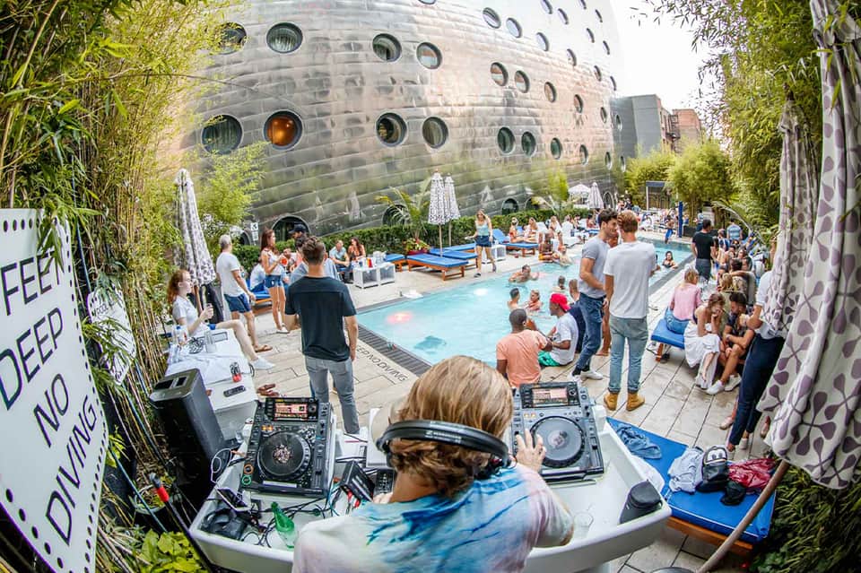 DJ performing at a lively poolside party with guests and a distinctive cylindrical building with circular windows