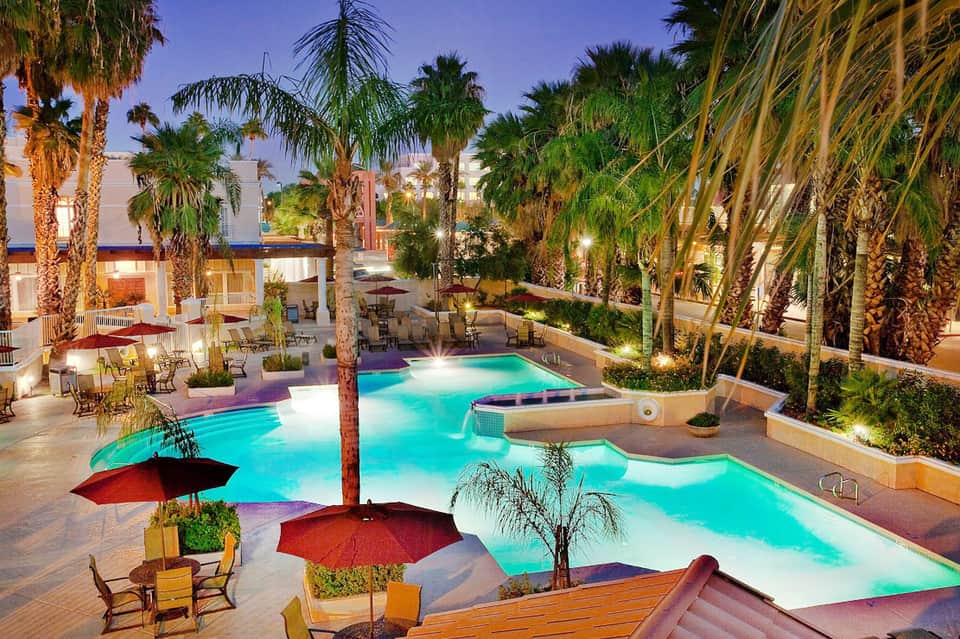 Illuminated resort pool complex surrounded by palm trees with lounge areas and tropical landscaping at dusk