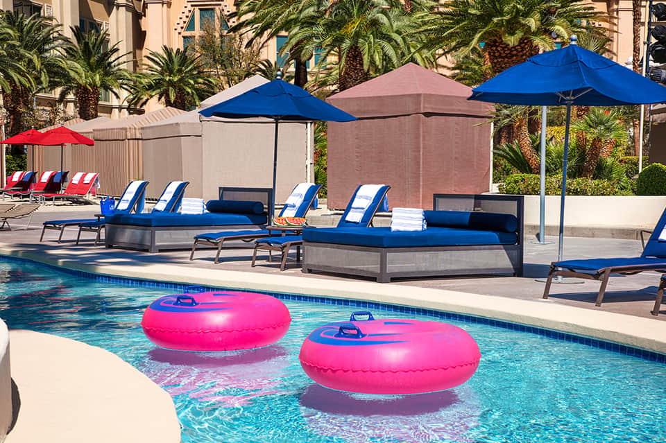 Pool area with blue lounge chairs, pink floats, and palm trees in sunny courtyard setting
