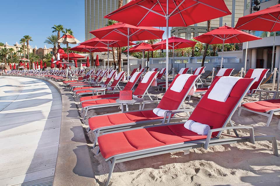 Beach club with red and white striped lounge chairs under red umbrellas, palm trees and high-rise buildings visible