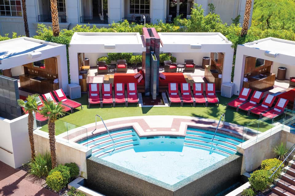 Outdoor hotel pool area with red loungers, white cabanas, and palm trees under sunny sky