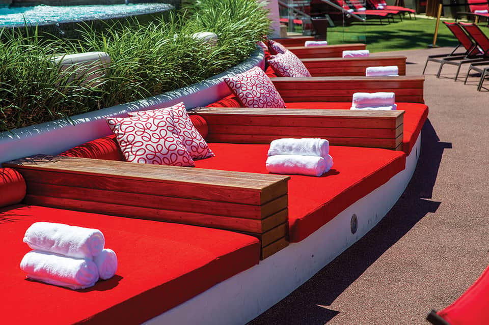 Red lounge beds with patterned pillows and rolled towels arranged along a poolside deck