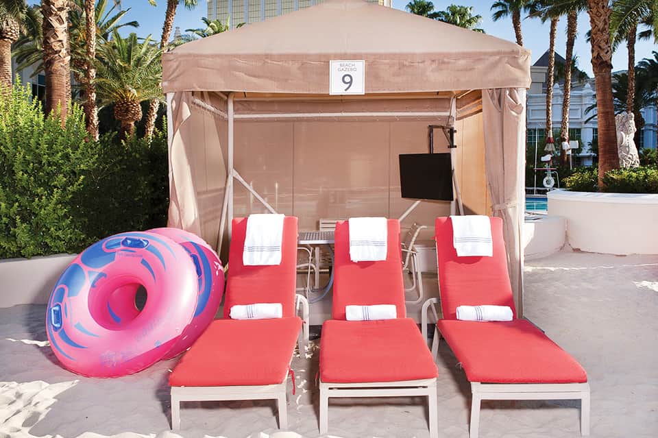 Beachside cabana with three red loungers, white towels, and pink float ring under tan tent numbered 9