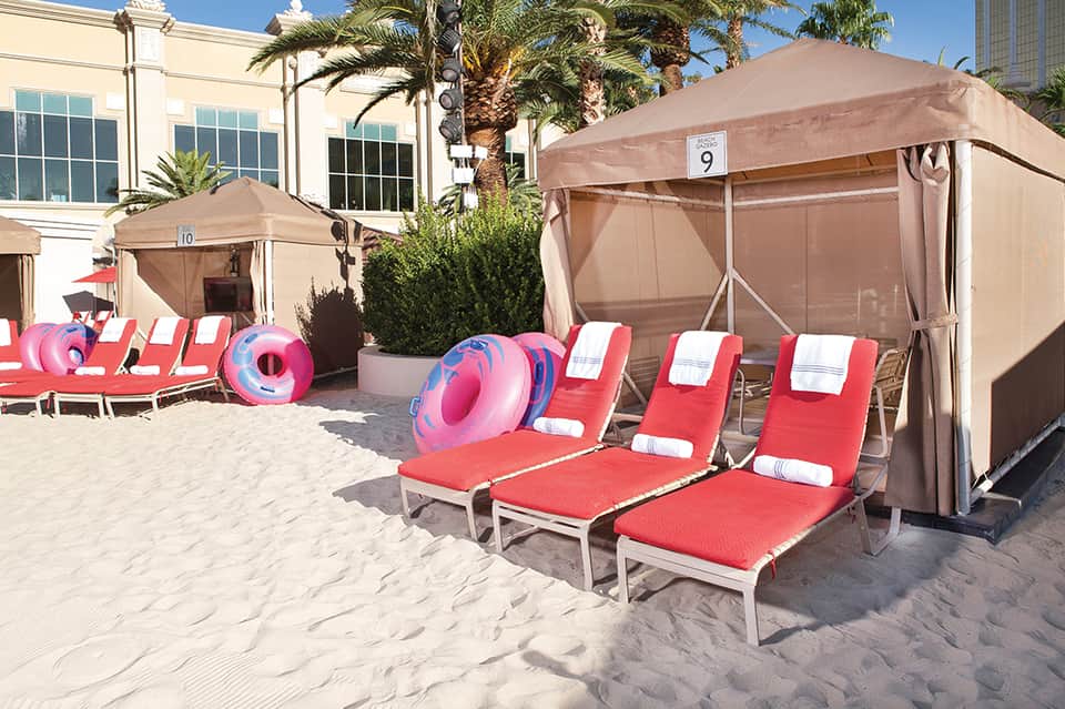 Beach cabanas with red loungers and pink floaties under tan umbrellas on sandy shore