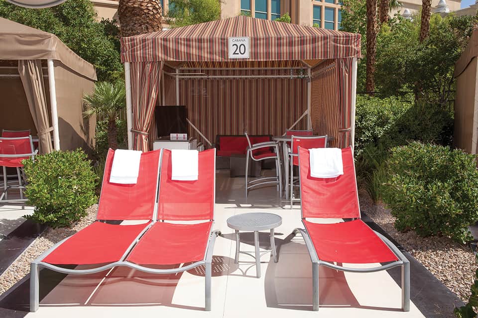 Poolside cabana area with red and white striped tent, red lounge chairs, small round table, and lush green landscaping