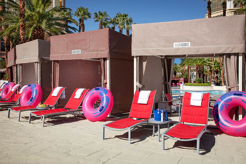 Beach cabanas with pink and blue floats arranged on sand with palm trees and pool area in background