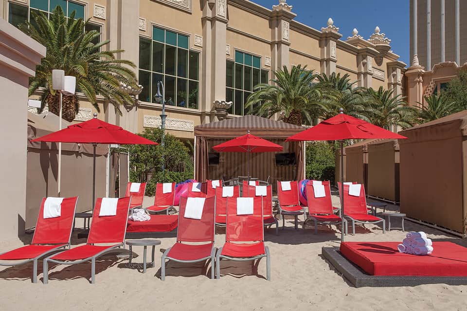Beach courtyard with red loungers, umbrellas, and palm trees surrounding a sandy area with modern hotel architecture
