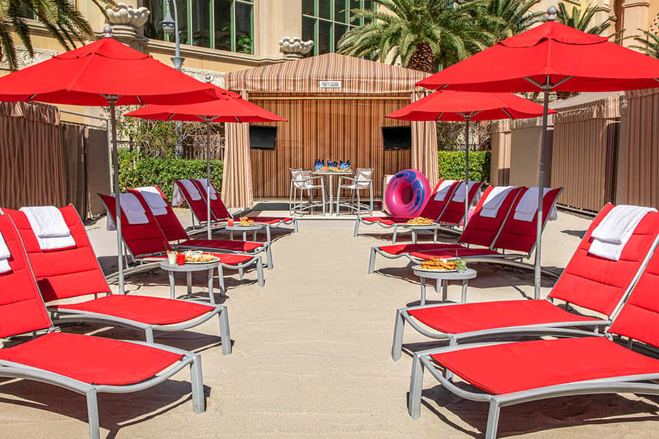 Outdoor pool deck with red loungers, umbrellas, and dining area surrounded by palm trees