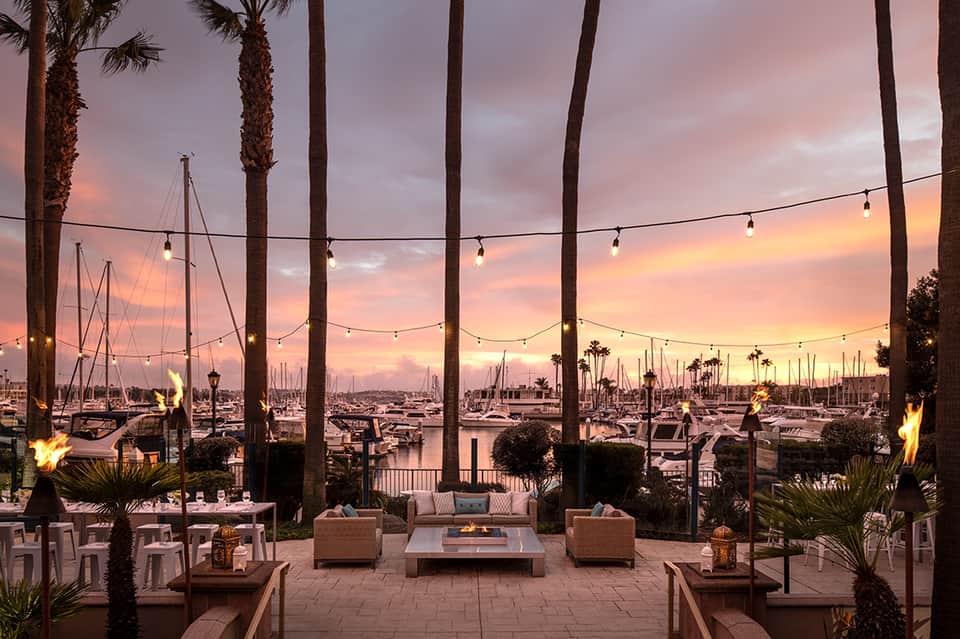 Waterfront terrace at sunset with palm trees, string lights, sailboats, and lounge seating overlooking marina