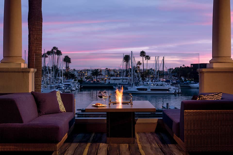 Waterfront lounge at sunset with fire pit, purple seating, and sailboats docked in marina background