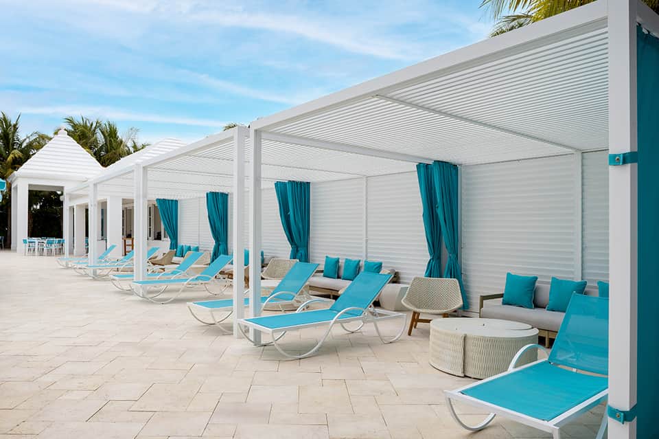 Modern poolside cabanas with turquoise loungers, white pergolas, and teal curtains along a light stone deck