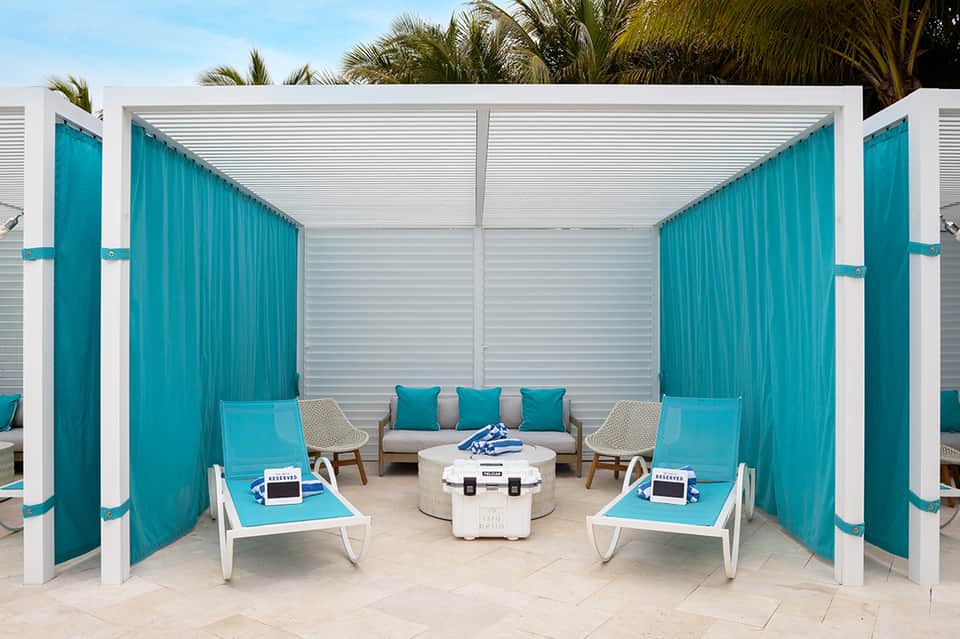 Luxury poolside cabana with turquoise curtains, white lounge chairs, and central seating area