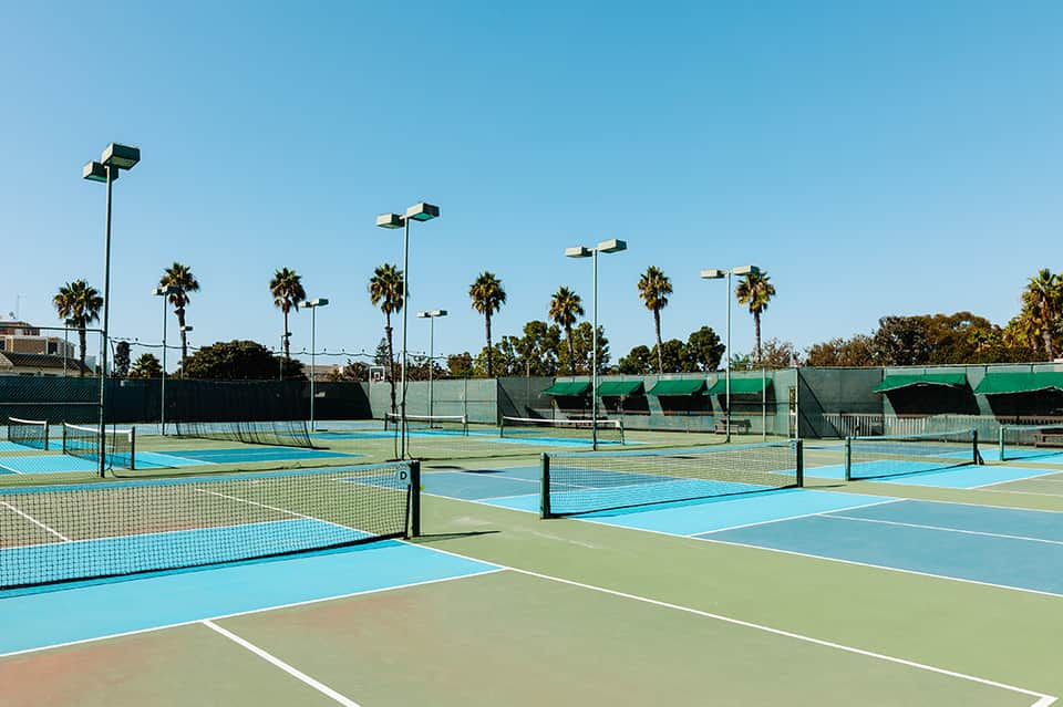 Outdoor tennis courts with blue and green playing surfaces, palm trees, and bright lighting poles under clear sky
