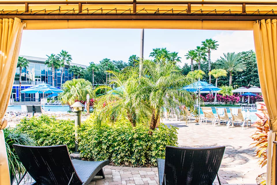 Resort pool area with palm trees, lounge chairs, and yellow cabanas viewed from covered patio