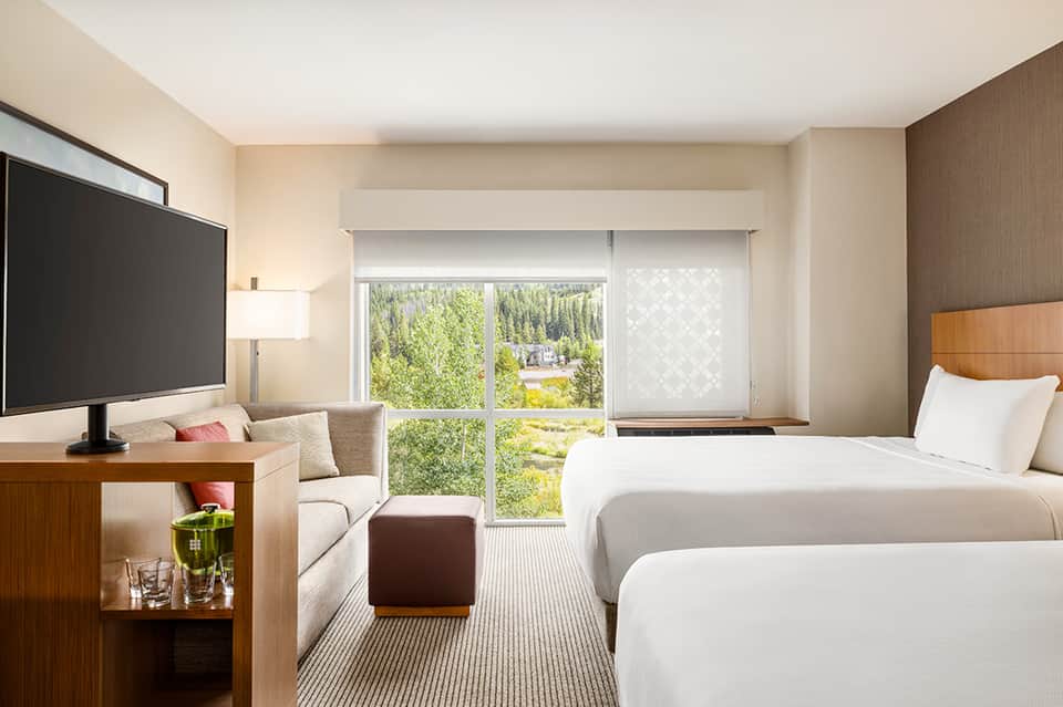 Modern hotel room with two queen beds, flat-screen TV, seating area, and floor-to-ceiling windows overlooking green landscape