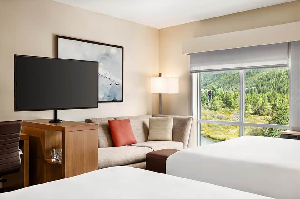 Modern hotel room with two beds, mountain forest view through glass doors, TV and seating area