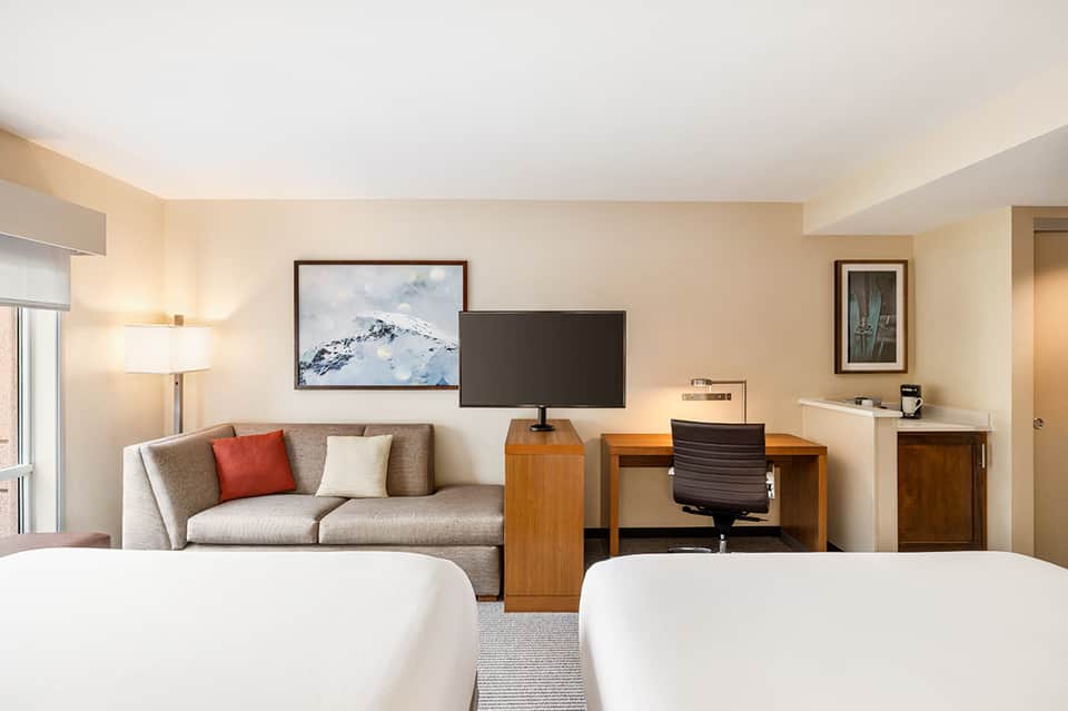 Modern hotel room with two queen beds, sofa, desk, TV, and mountain landscape artwork
