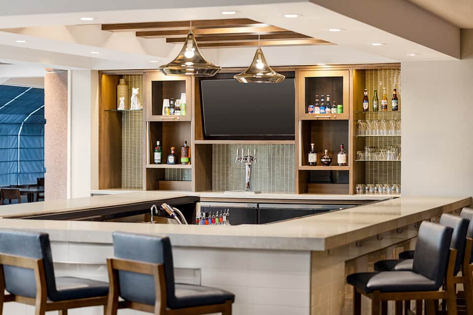 Modern hotel bar with wooden shelving, brass pendant lights, and a curved white counter with bar seating