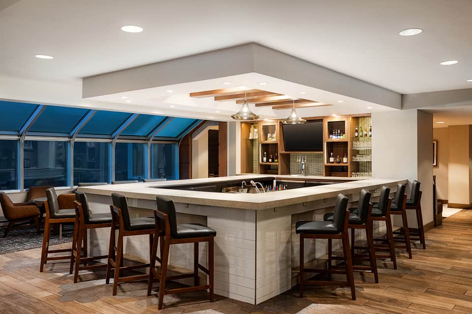 Upscale indoor bar with white stone counter, wooden bar stools, blue-tinted windows, and ambient lighting fixtures