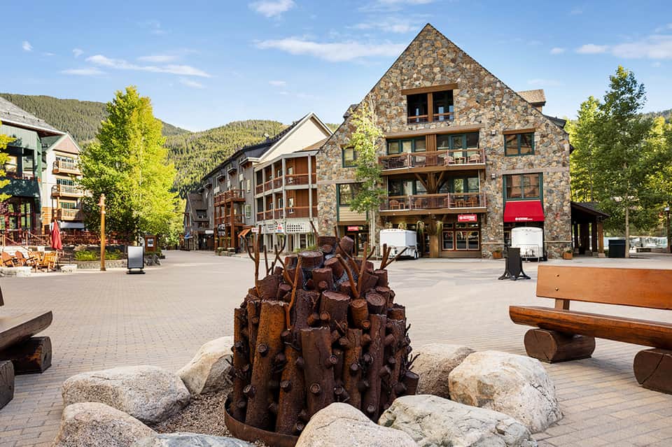 Mountain village plaza with stone buildings, wooden benches, and carved wooden sculpture surrounded by evergreen trees