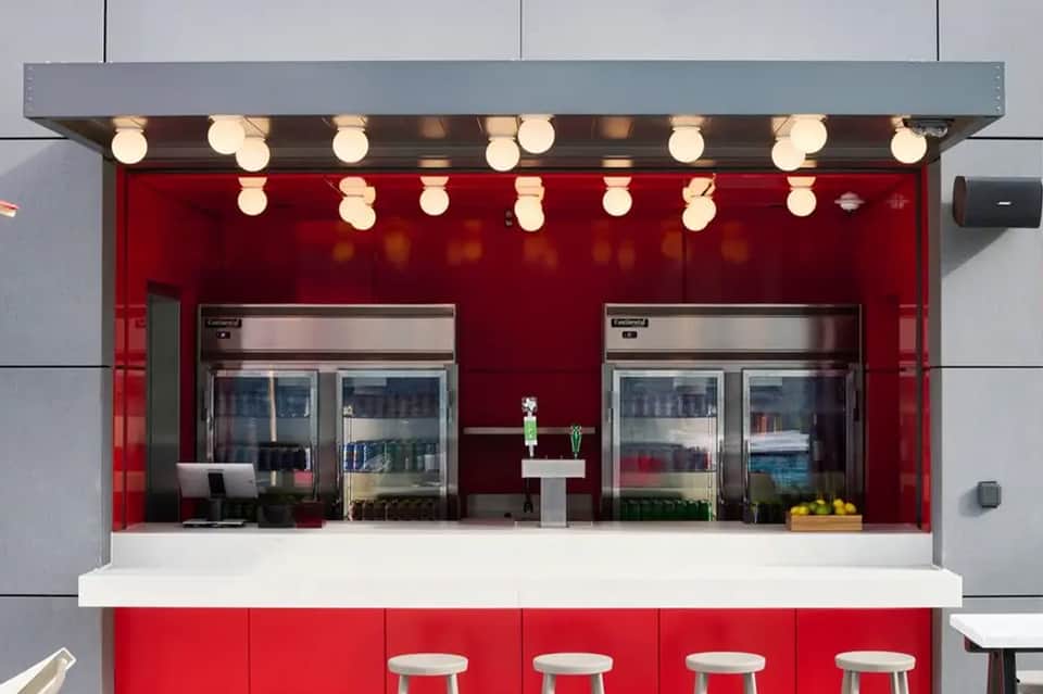 Modern red food kiosk with illuminated counter, refrigerated cases, and white seating stools