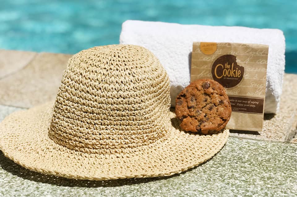 Straw hat, chocolate chip cookie, and white towel arranged on sandy beach with turquoise ocean background