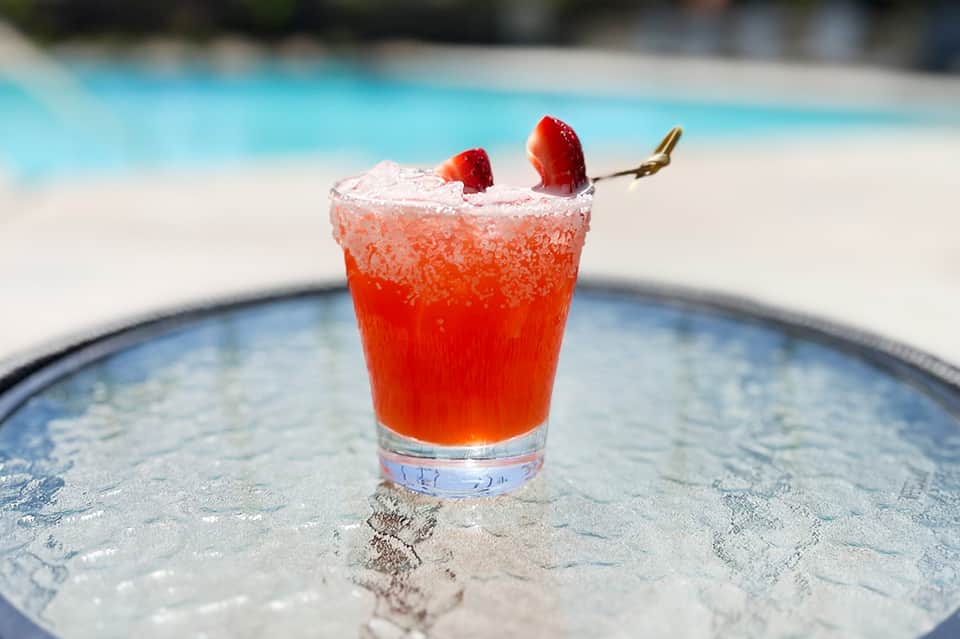 Red cocktail with sugar rim and strawberry garnish on poolside table overlooking blue water