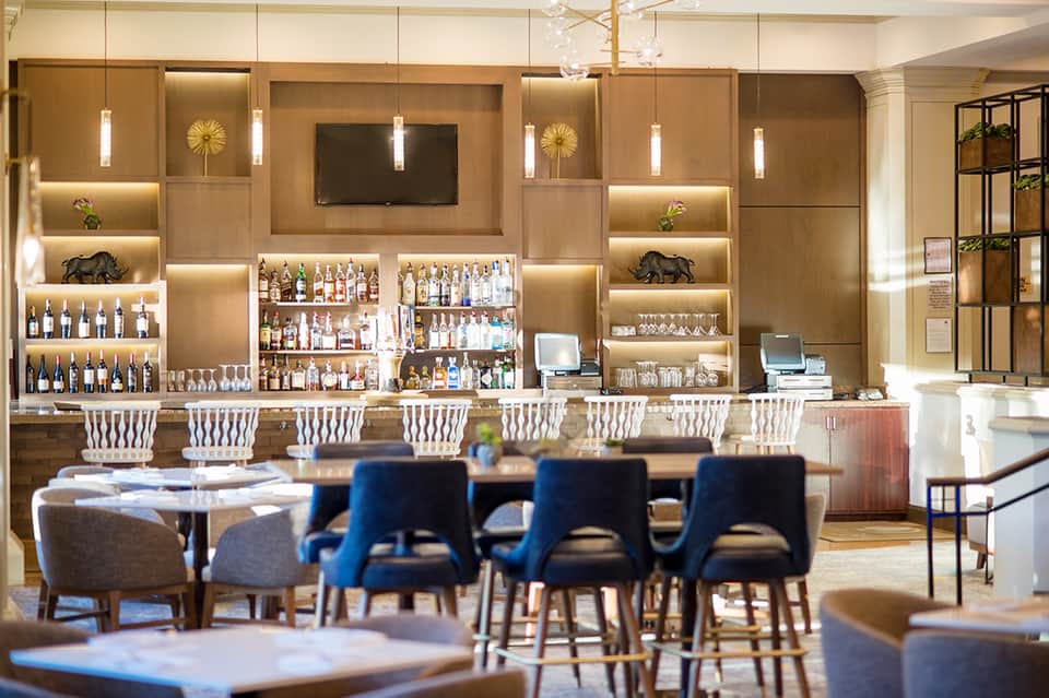 Modern hotel bar with illuminated shelving, white bar seating, wooden accents, and contemporary pendant lighting