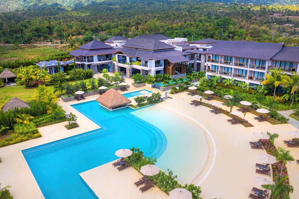 Aerial view of luxury resort with curved blue pool, beige beach area, and tropical landscaping surrounded by lush green forest
