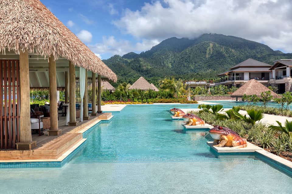 Luxury resort pool with turquoise water, thatched-roof pavilion, and lush mountain backdrop