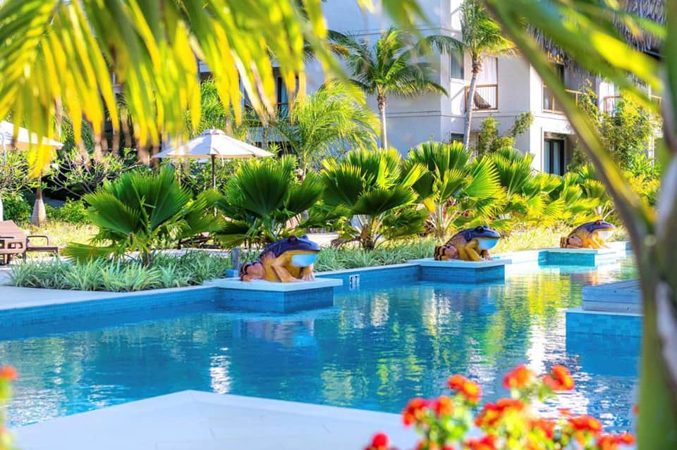 Tropical resort pool with blue water, palm trees, and guests lounging on deck platforms