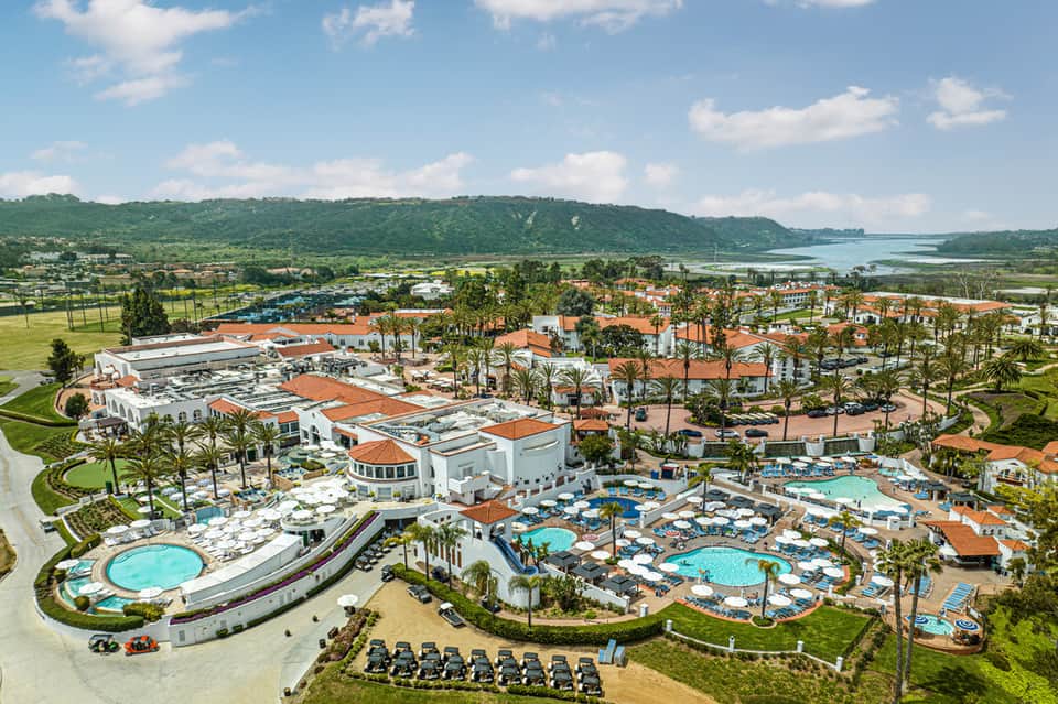 Aerial view of a sprawling resort with pools, palm trees, orange-roofed buildings, and lake views