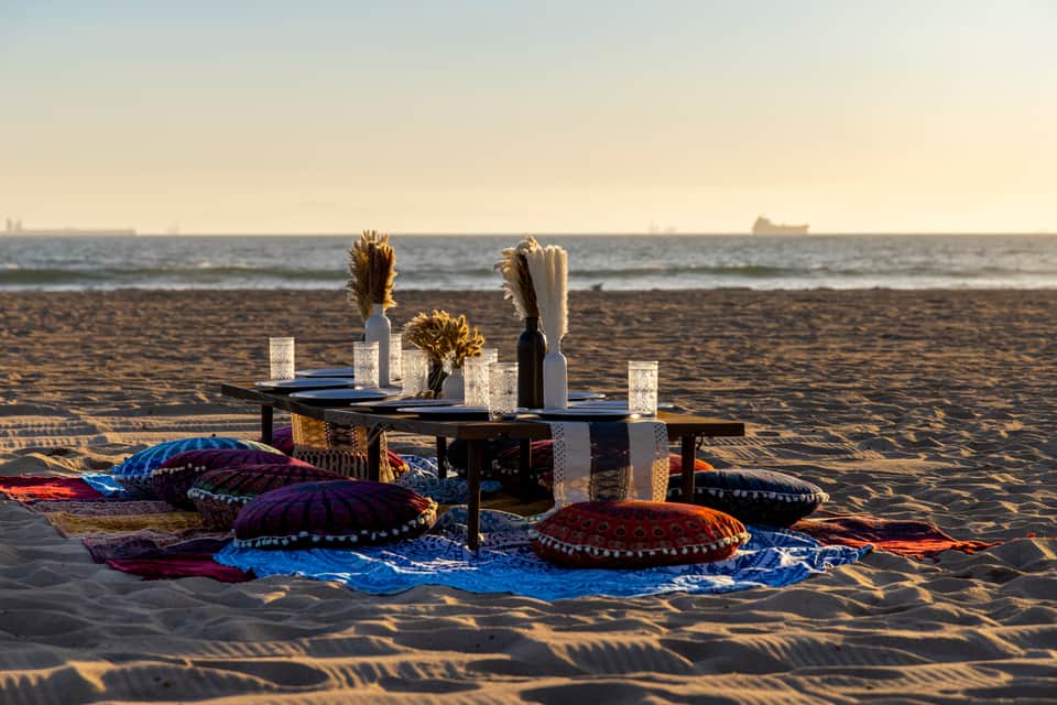 Beachfront dinner setup with colorful patterned cushions, candles, and floral centerpieces on sand at sunset