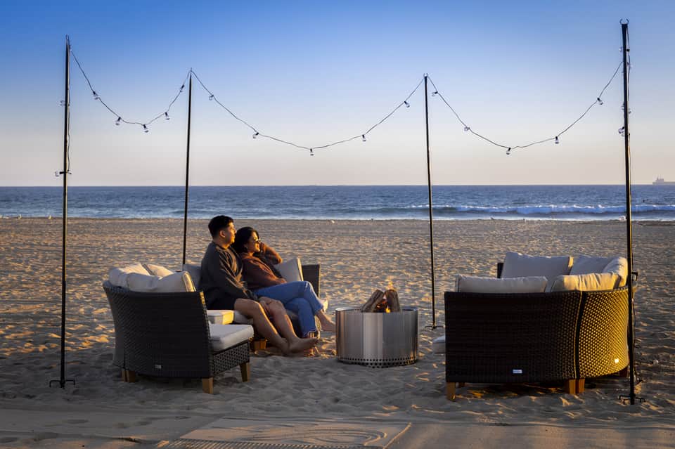 Couple relaxing in wicker lounge chairs on beach at sunset with strung lights overhead