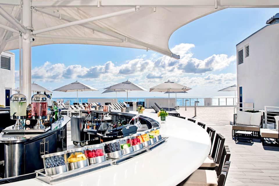 Rooftop bar with white umbrellas, bar counter with colorful garnishes, and ocean views under blue sky