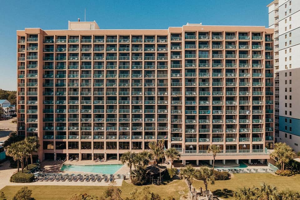 Beachfront resort building with coral-colored brick facade, multiple balconies, and oceanside pool surrounded by palm trees