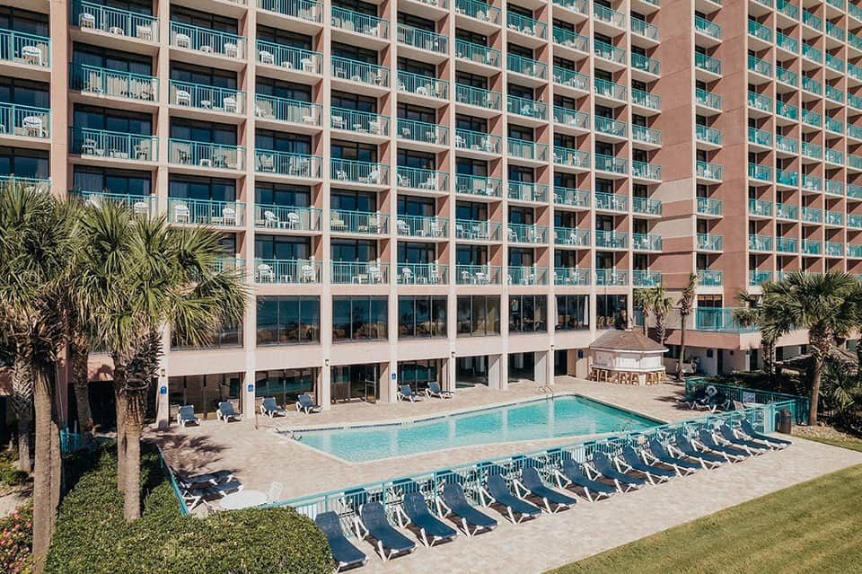 Large beachfront resort with red brick building, balconies, and outdoor pool with lounge chairs