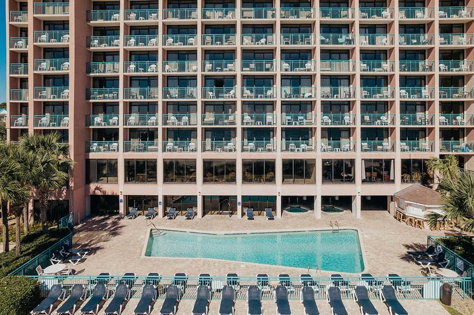 Multi-story beachfront hotel with turquoise pool, lounge chairs, and palm trees on sandy beach