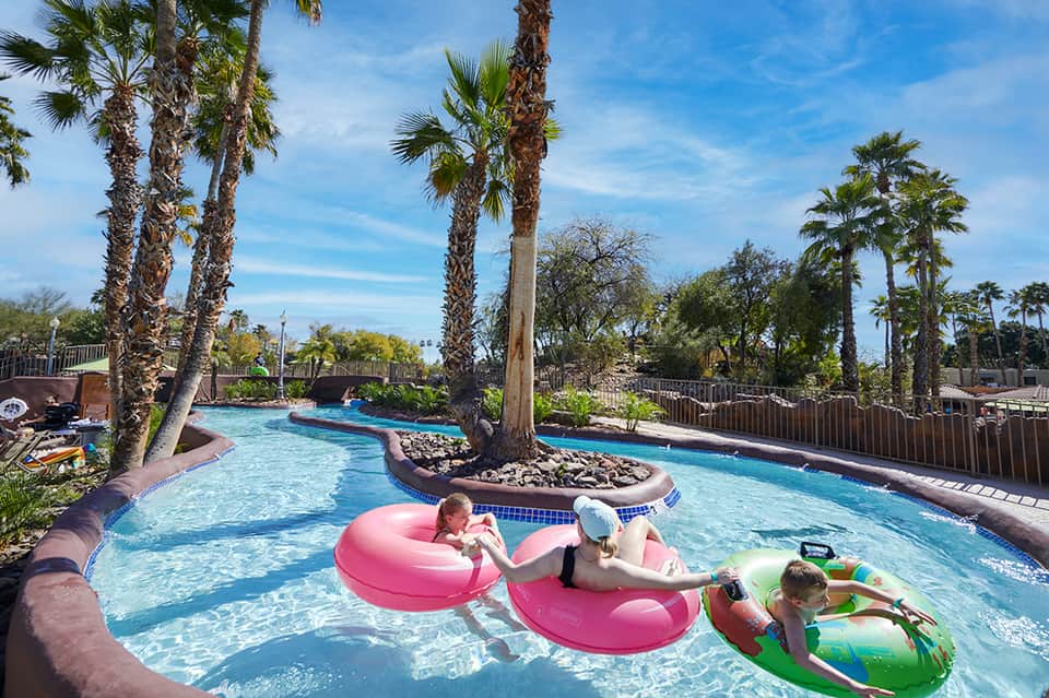 Lazy river with colorful inflatable tubes and children playing, surrounded by palm trees and blue sky