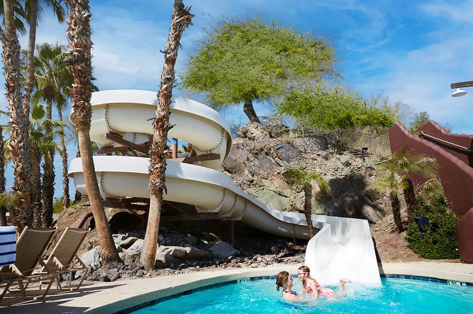 Spiral white water slide at resort pool surrounded by palm trees and lounge chairs