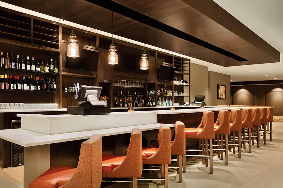 Modern hotel bar with dark wood cabinetry, white marble countertop, pendant lighting, and orange upholstered bar stools