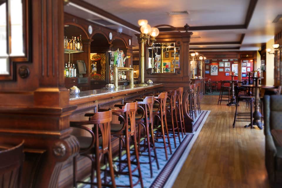 Upscale pub interior with dark wood bar, vintage lighting, brass fixtures, and wooden bar stools along counter