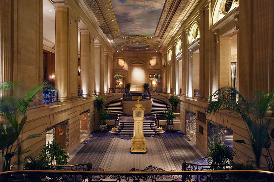Grand classical hallway with ornate columns, decorative ceiling, potted plants, and marble floors