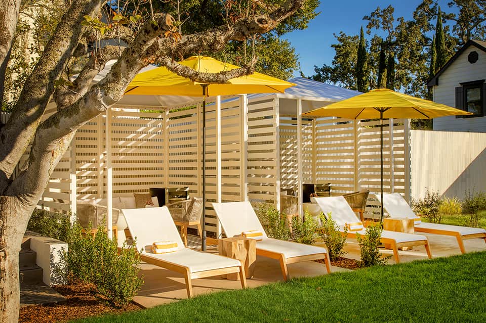 Backyard lounge area with wooden sun loungers, yellow umbrellas, and slatted privacy screens under mature trees