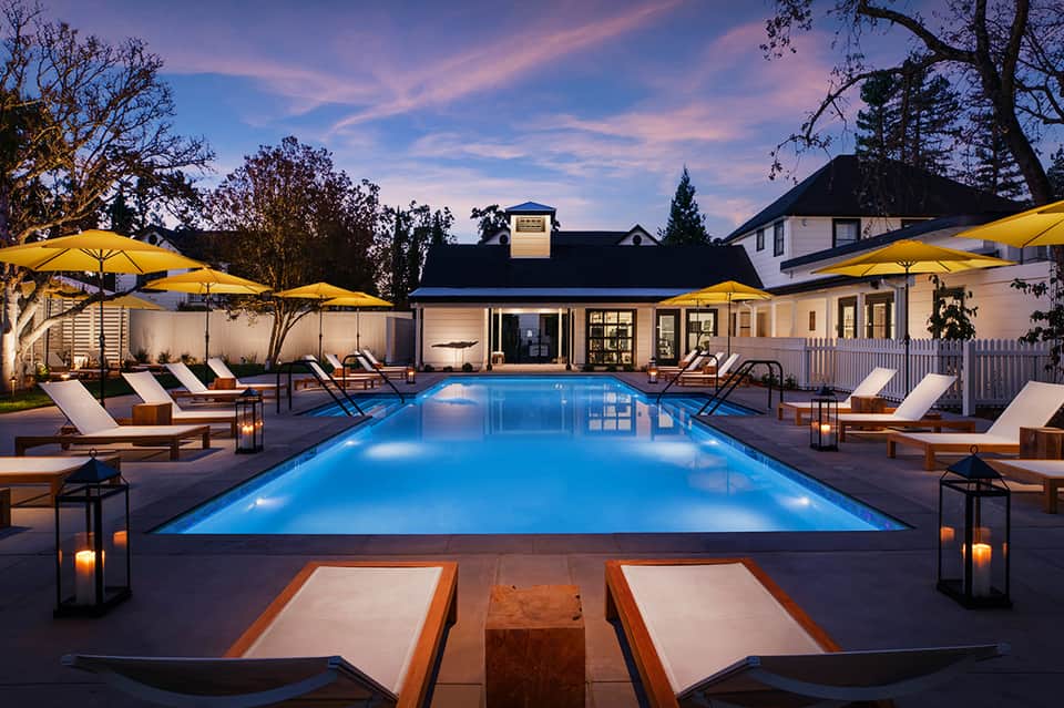 Illuminated rectangular pool at dusk with yellow umbrellas, lounge chairs, and modern white buildings