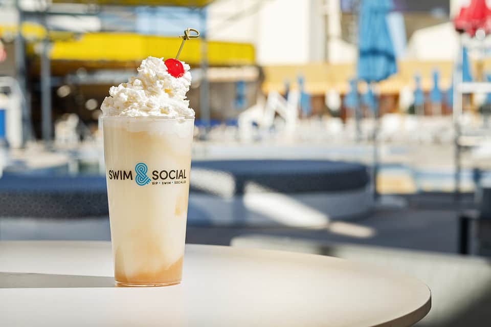 Whipped cream topped beverage with cherry garnish on Swim & Social branded cup by poolside