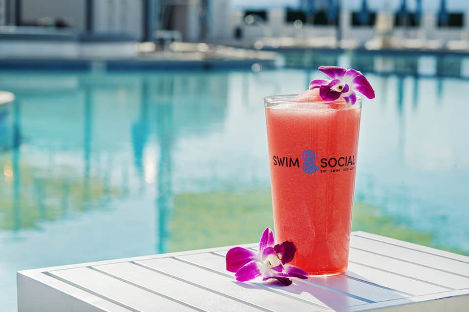 Coral cocktail with orchid flower garnish at poolside bar with turquoise water background